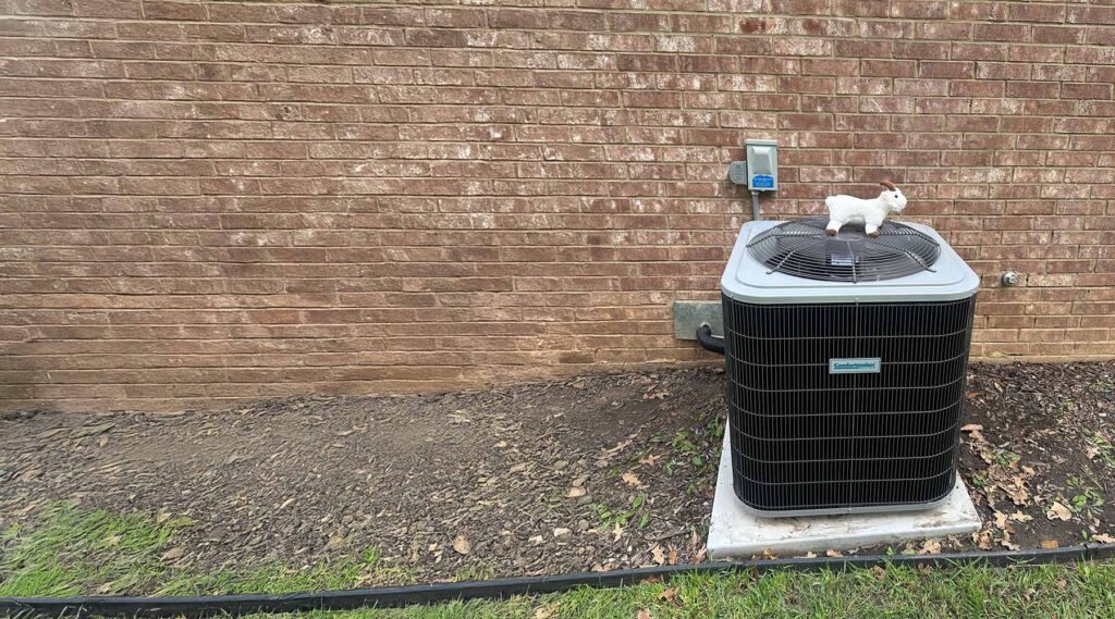 Air conditioner unit with a toy goat on top against a brick wall.