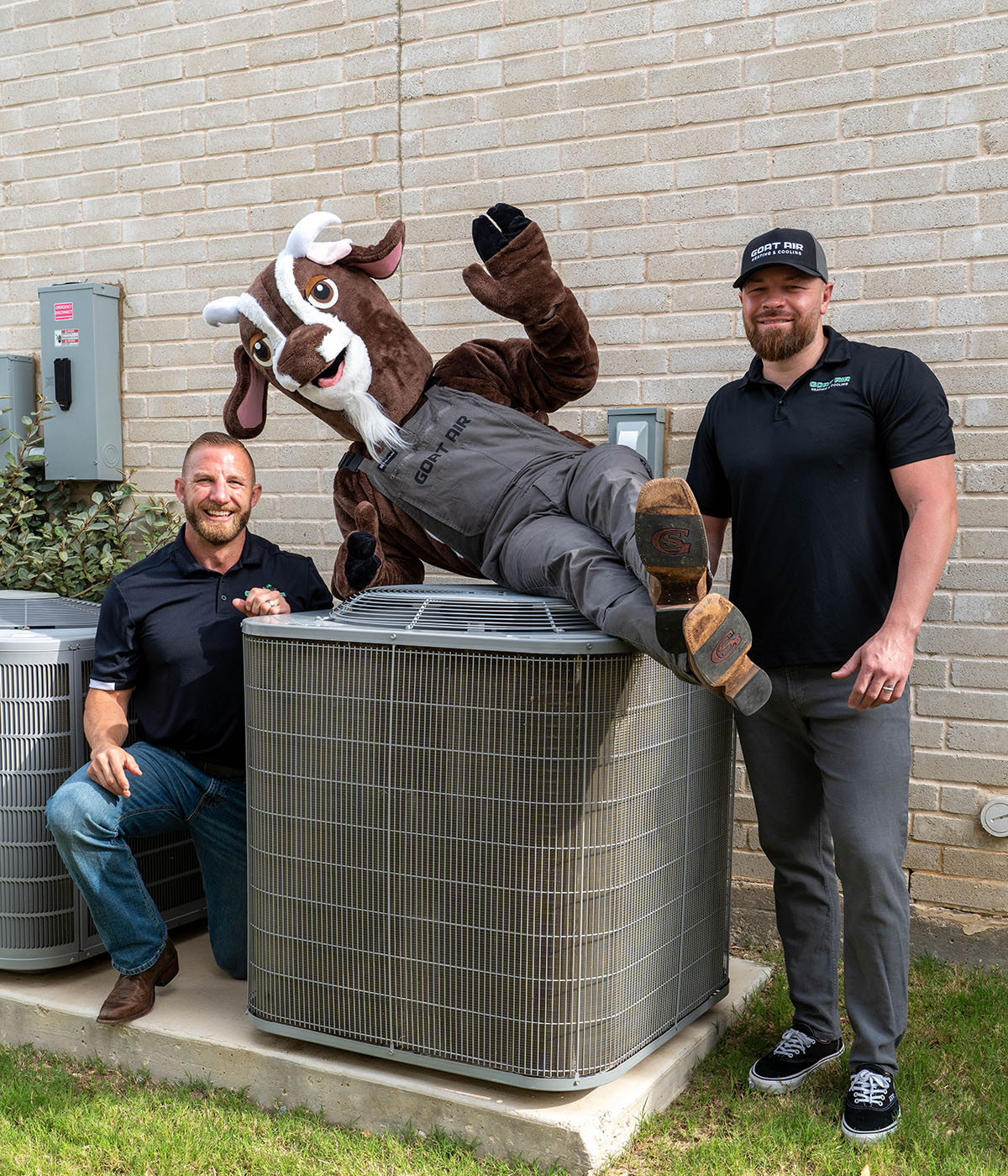 Two smiling Goat Air owners posing with the Goat Air costumed mascot playfully flopped over an outdoor AC condenser unit beside multiple units along a brick wall.
