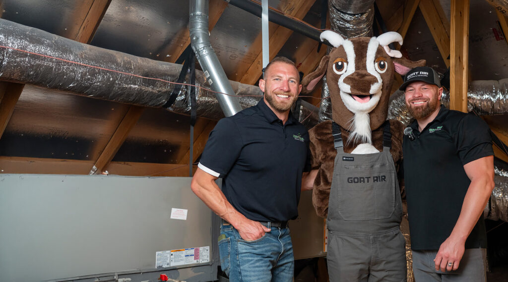 Two smiling Goat Air Heating & Cooling owners in black branded polos posing with the Goat Air costumed mascot inside a home attic next to an air handler unit and flexible ductwork.