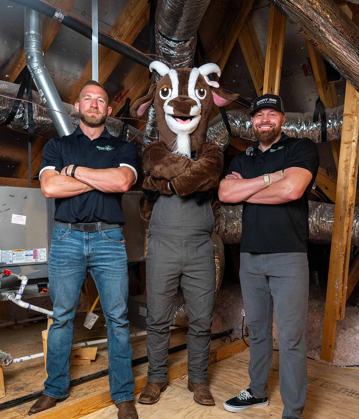 The two Goat Air owners standing with arms crossed on either side of the Goat Air costumed mascot inside a home attic next to an air handler unit and flexible ductwork.