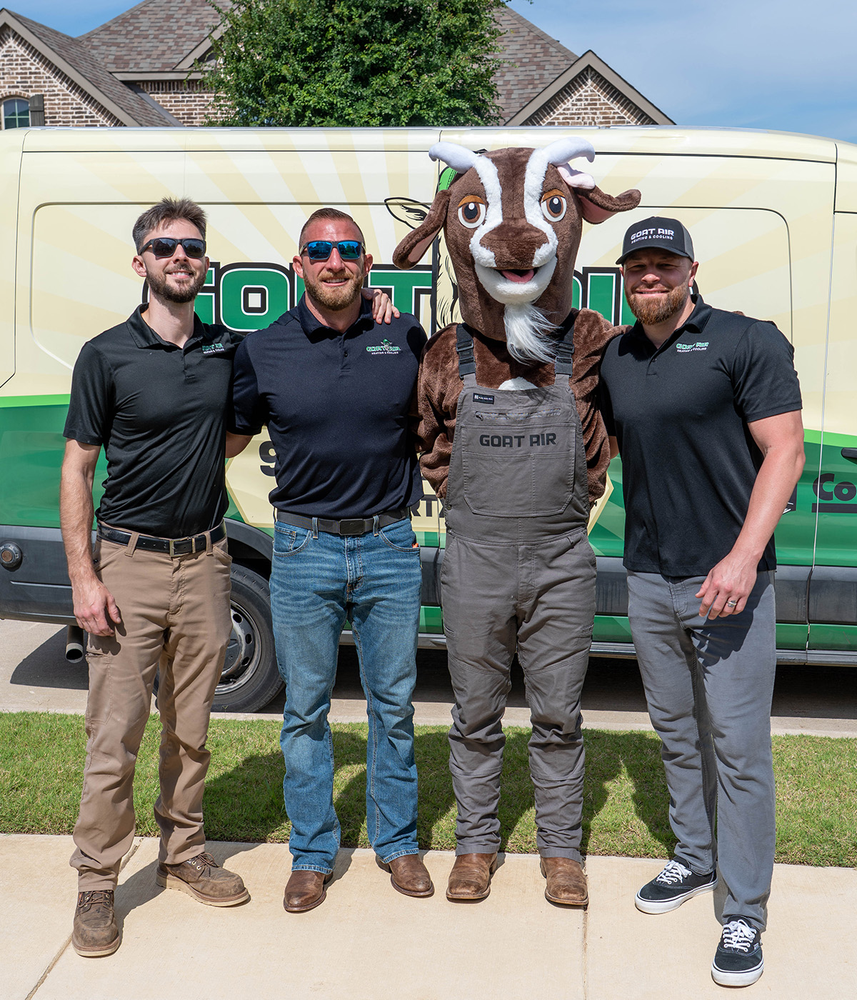 Three Goat Air Heating & Cooling team members in black branded polos posing with the Goat Air costumed mascot character in overalls in front of their service van in a North Texas neighborhood.