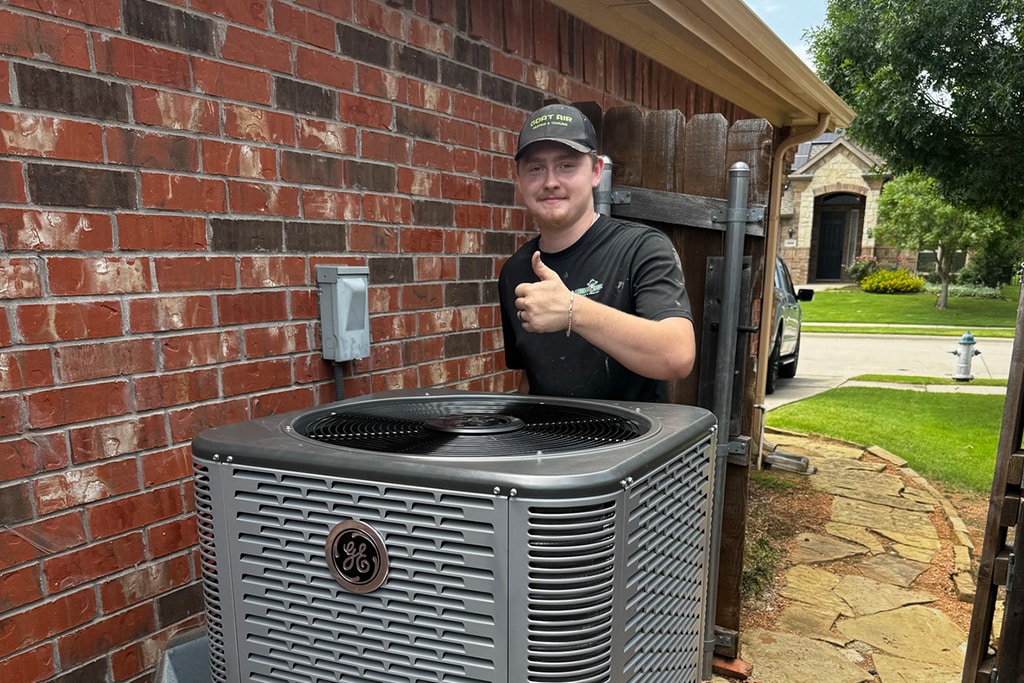 Goat Air technician giving a thumbs up beside a newly installed GE central air conditioning unit outside a brick home